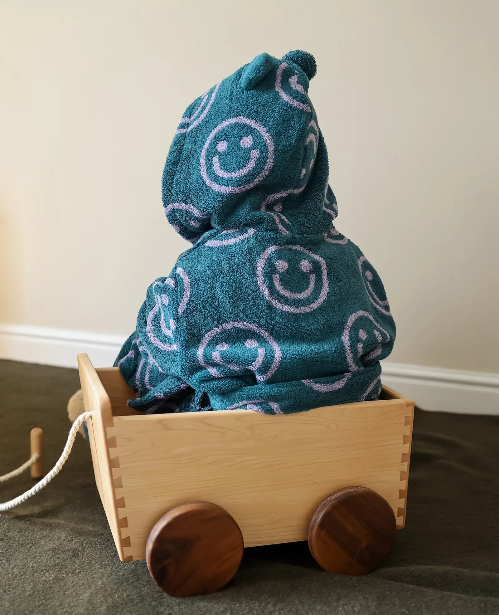 Back view of a child sitting in a wooden cart showing the purple smiley face pattern on the teal hooded robe