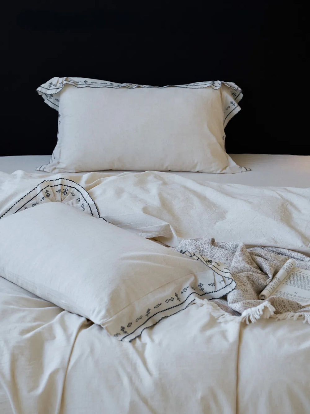Close-up of Textured Embroidered Ruffle Linen Pillowcases on a neatly made bed against a black wall - RoomDen.