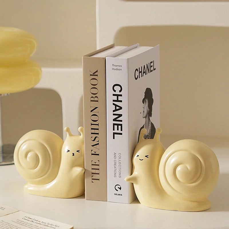 A pair of cute pastel yellow smiley snail ceramic bookends holding up fashion books on a white desk - RoomDen.