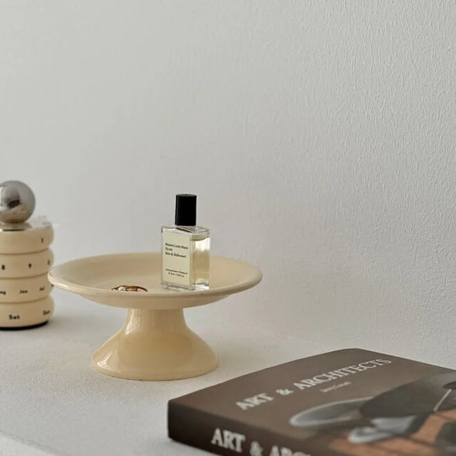 A creamy beige pedestal jewelry dish on a white shelf, holding a perfume bottle and rings, next to an art book. - RoomDen