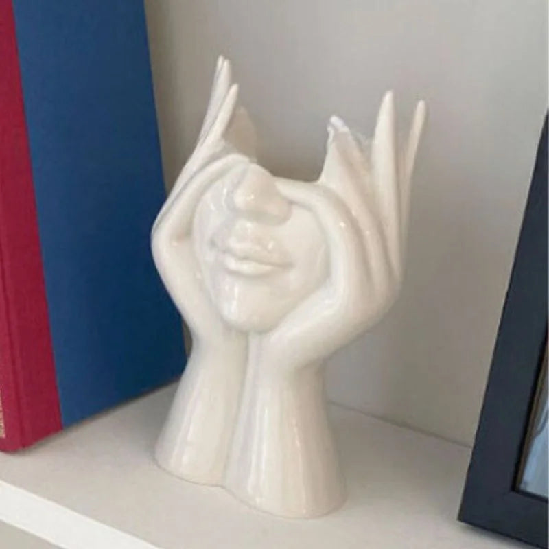 Alternate 'Peekaboo' design of the white sculptural face vase with hands on cheeks, on a bookshelf - RoomDen.