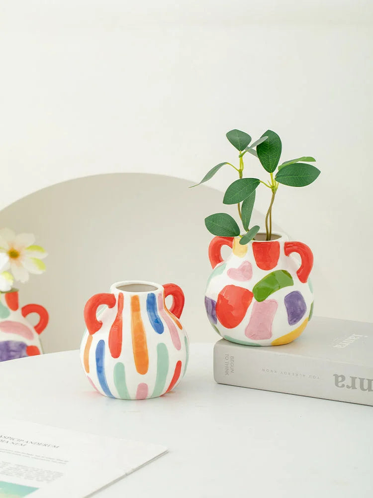 Two colorful abstract ceramic vases, one striped and one spotty, styled on a white table with a plant and books. - RoomDen