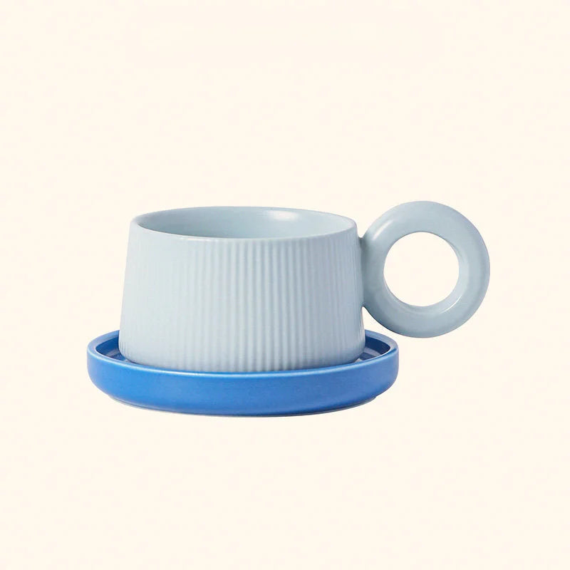 Front view of the Sky Blue ribbed ceramic mug with its round donut handle, sitting on a royal blue saucer. - RoomDen