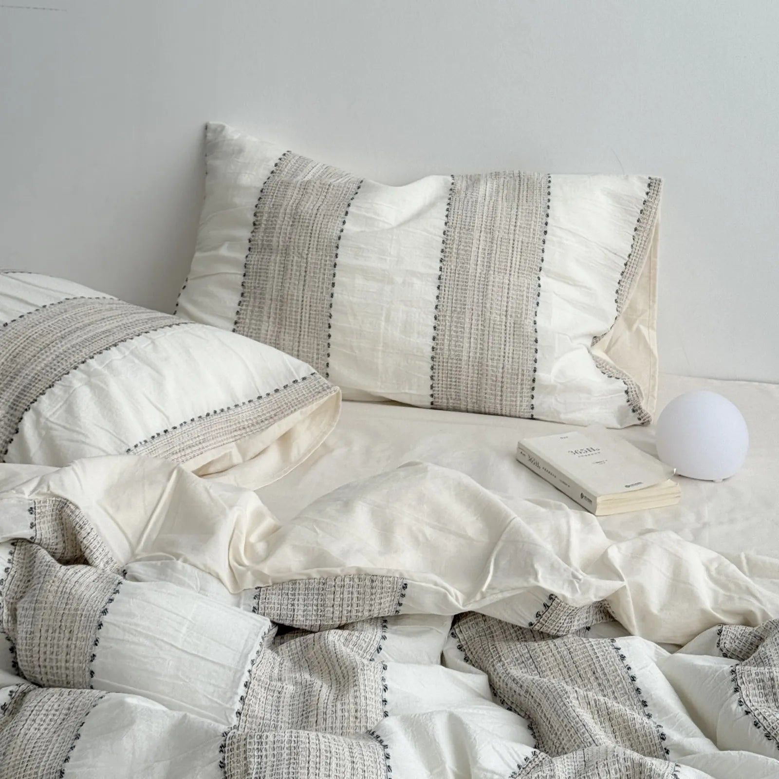 Full view of the Oatmeal Beige woven stripe bedding set with a small white lamp and book on the bedside. - RoomDen