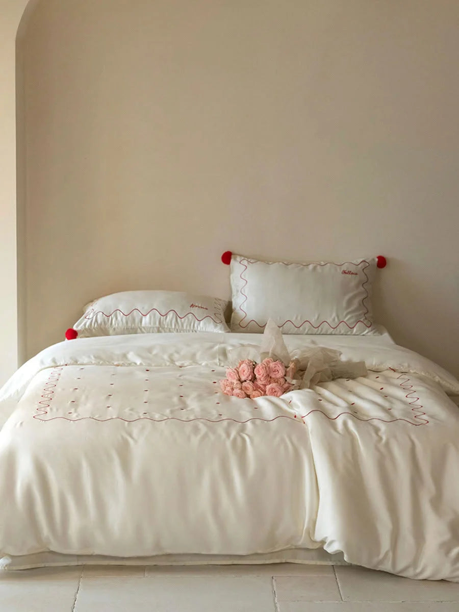 Full view of a bed made with the Creamy White & Red pom pom Tencel bedding set, with a bouquet of pink roses on top. - RoomDen
