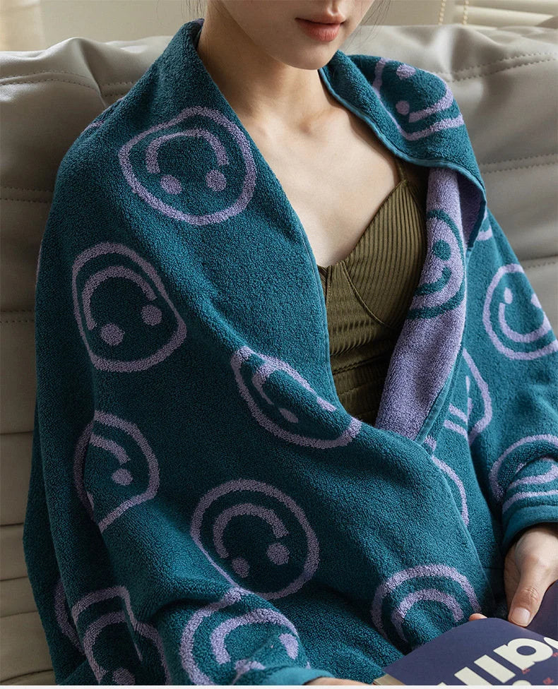 A person wrapped comfortably in the large, plush teal and lavender smiley face cotton bath towel, showcasing its size and softness - RoomDen.