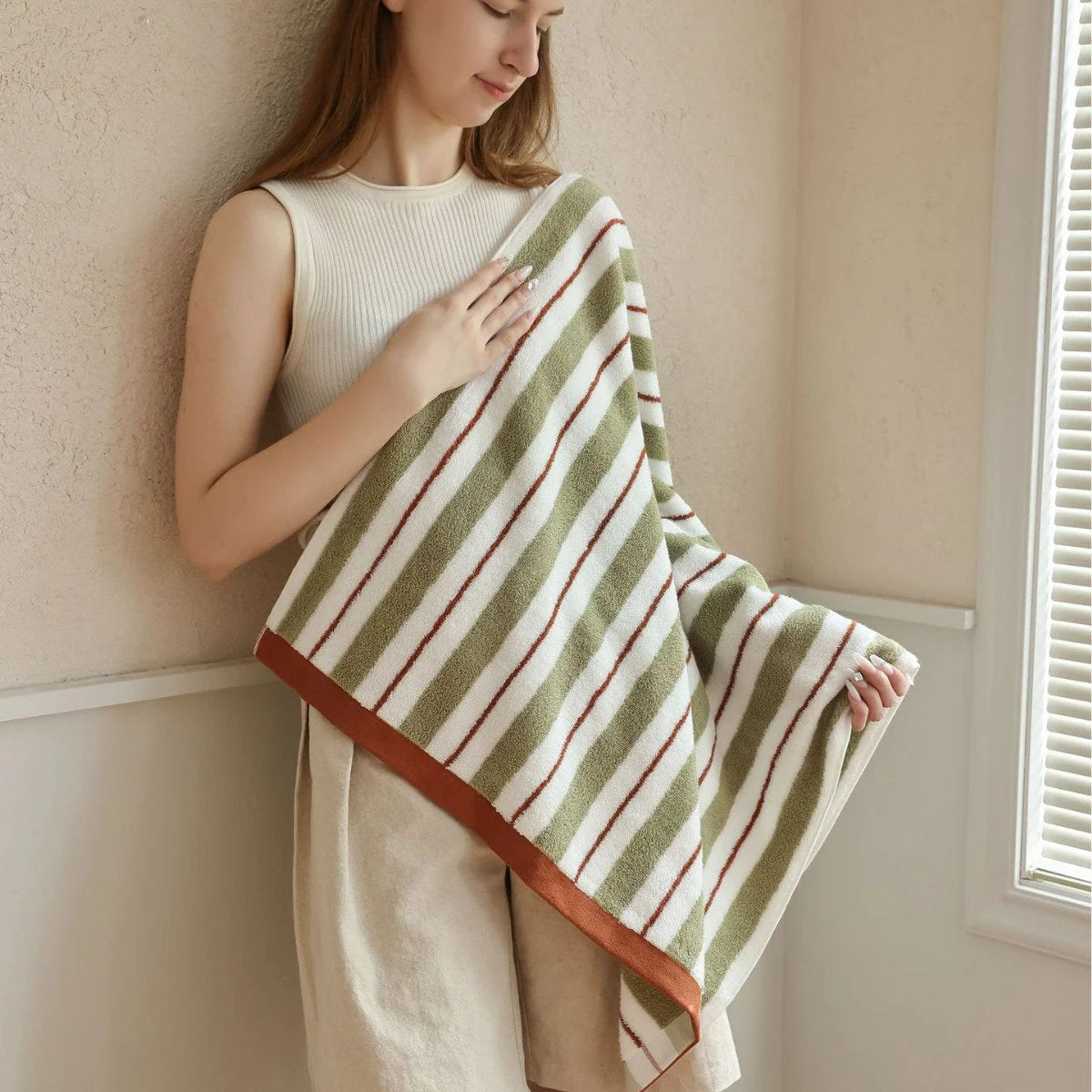 A woman holding the green and white striped cotton face towel against her shoulder in a softly lit room - RoomDen