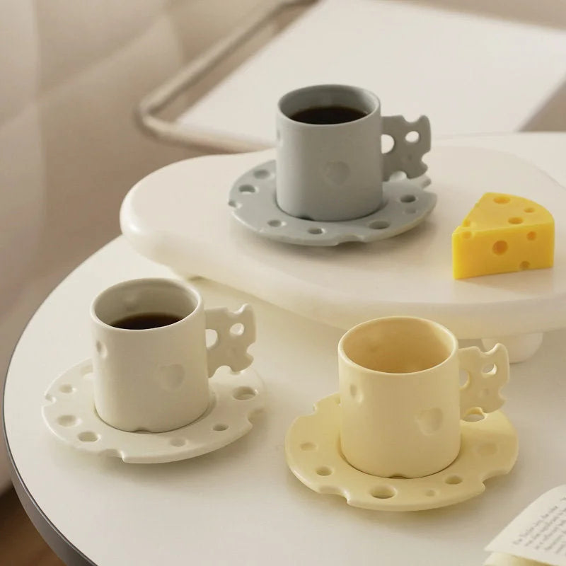 Three Cheese Ceramic Mug & Saucer sets in cream white, yellow, and light grey arranged on a table. - RoomDen