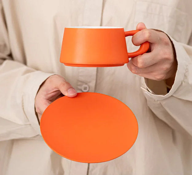 Close-up of hands holding a Marigold Orange ceramic coffee cup and its matching saucer. - RoomDen