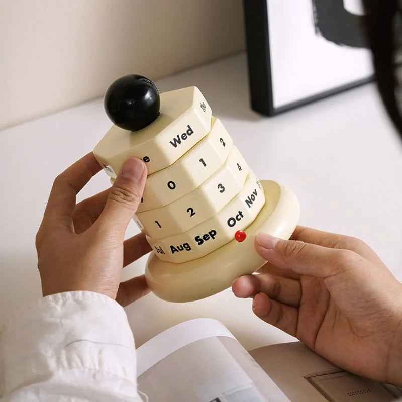 Hands holding and adjusting a creamy beige wooden perpetual calendar on a white desk. - RoomDen