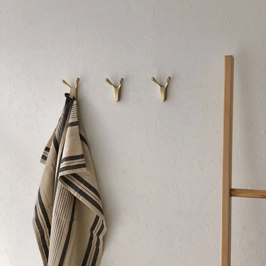 Three gold branch-shaped metal wall hooks mounted on a textured white wall, with a striped towel hanging from one - RoomDen