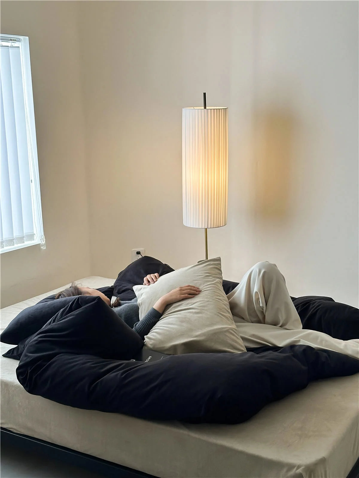 Person relaxing in a bed with an Obsidian Black & Cream velvet bedding set, next to a modern floor lamp. - RoomDen