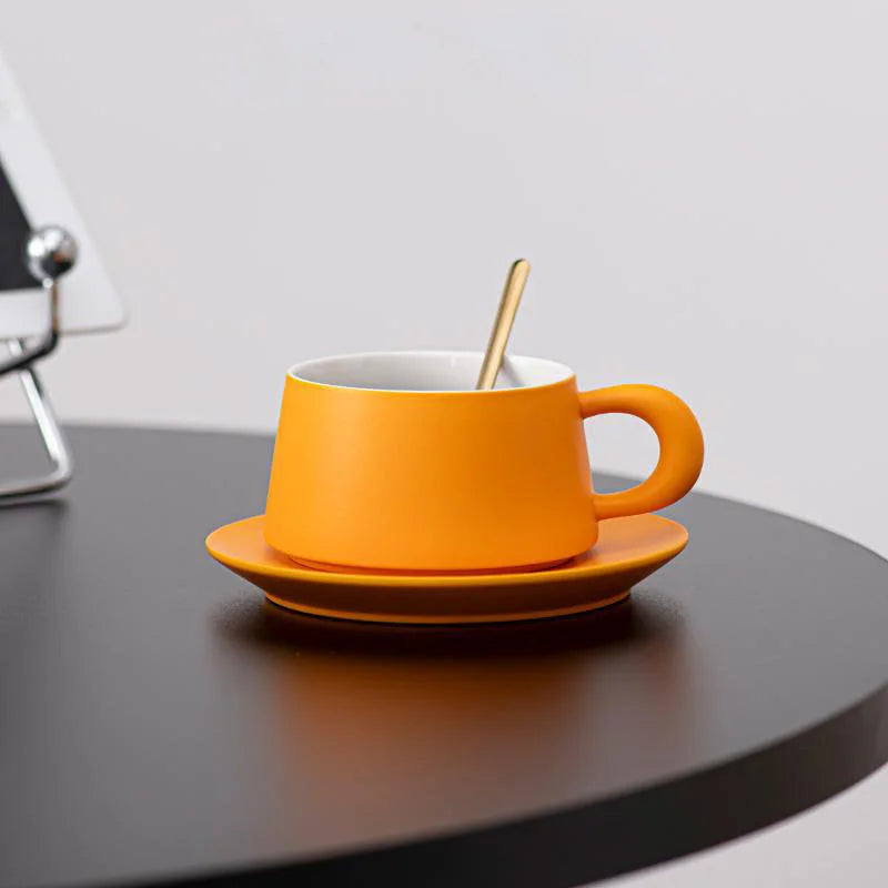 The Marigold Orange coffee cup and saucer set sitting on the edge of a modern black table. - RoomDen