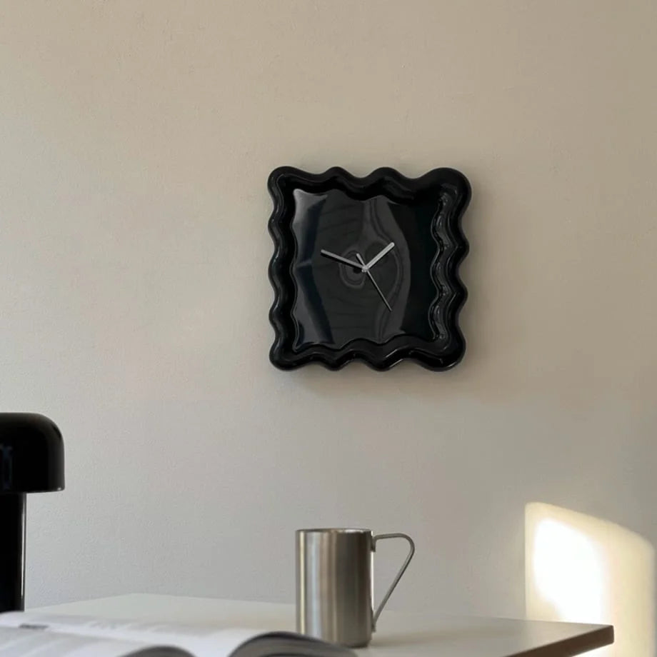 A black Wavy Squiggle Frame Clock mounted on a minimalist off-white wall above a desk. - RoomDen
