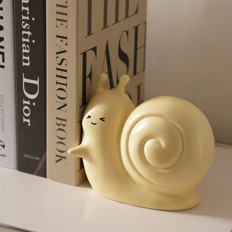 Detailed view of the pastel yellow snail bookend with a cute winking face, pushing against a stack of books - RoomDen.