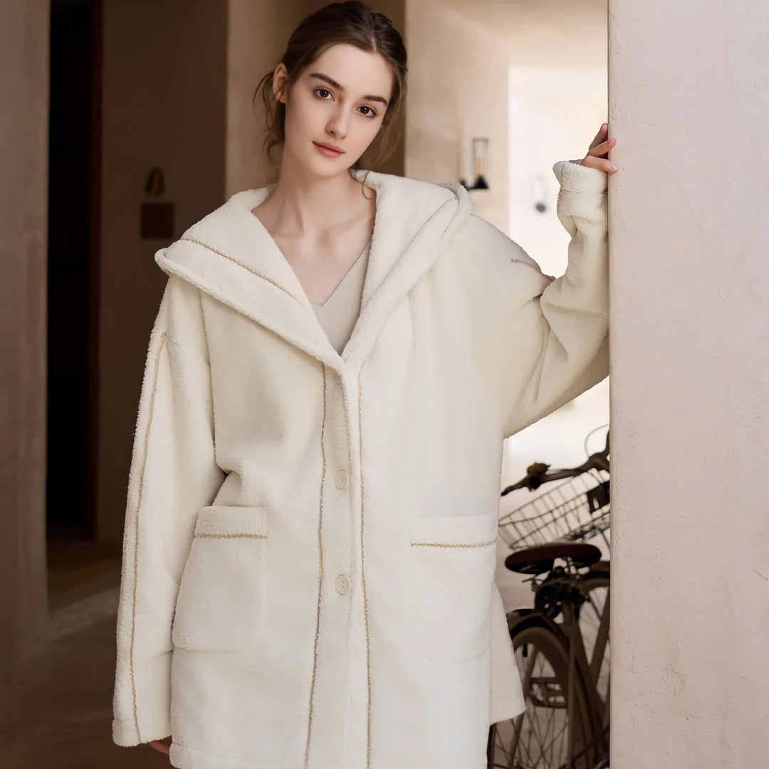 Model standing in a hallway wearing an unbelted creamy white hooded fleece pajama jacket with matching pants - RoomDen.