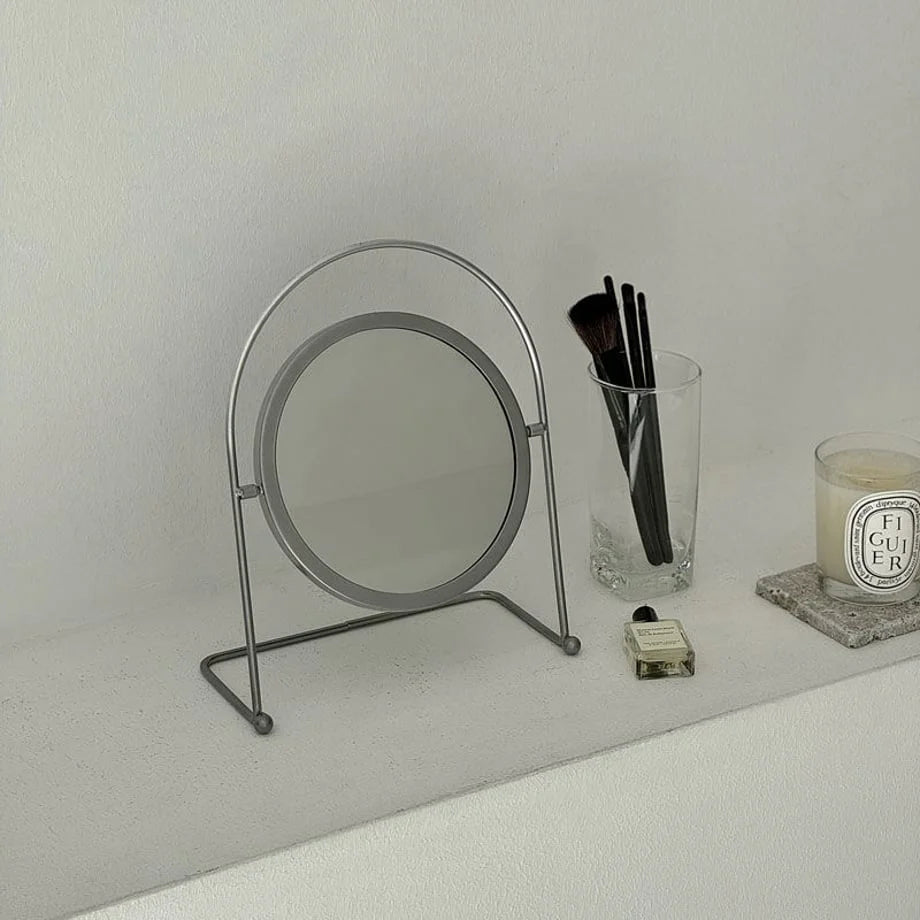 Front view of the round metal vanity mirror on a neutral-toned surface with accessories. - RoomDen