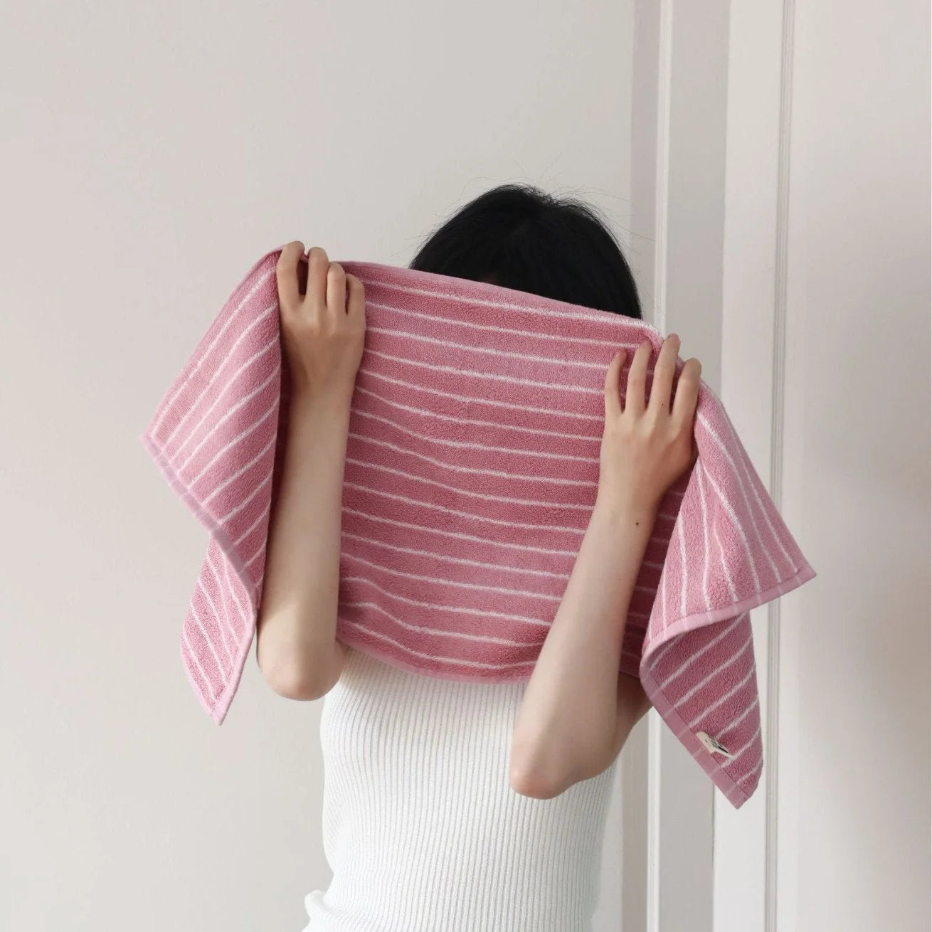 A woman gently drying her face with a plush pink and white pinstripe cotton face towel - RoomDen.