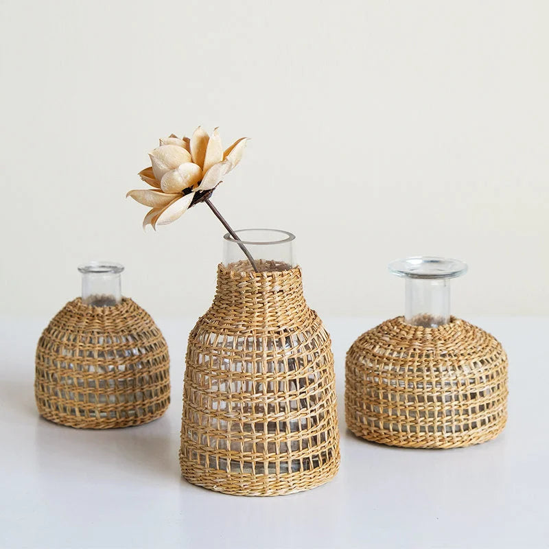 Lifestyle image showing the set of three Handwoven Rattan & Glass Bud Vases on a white surface, one with a dried flower.