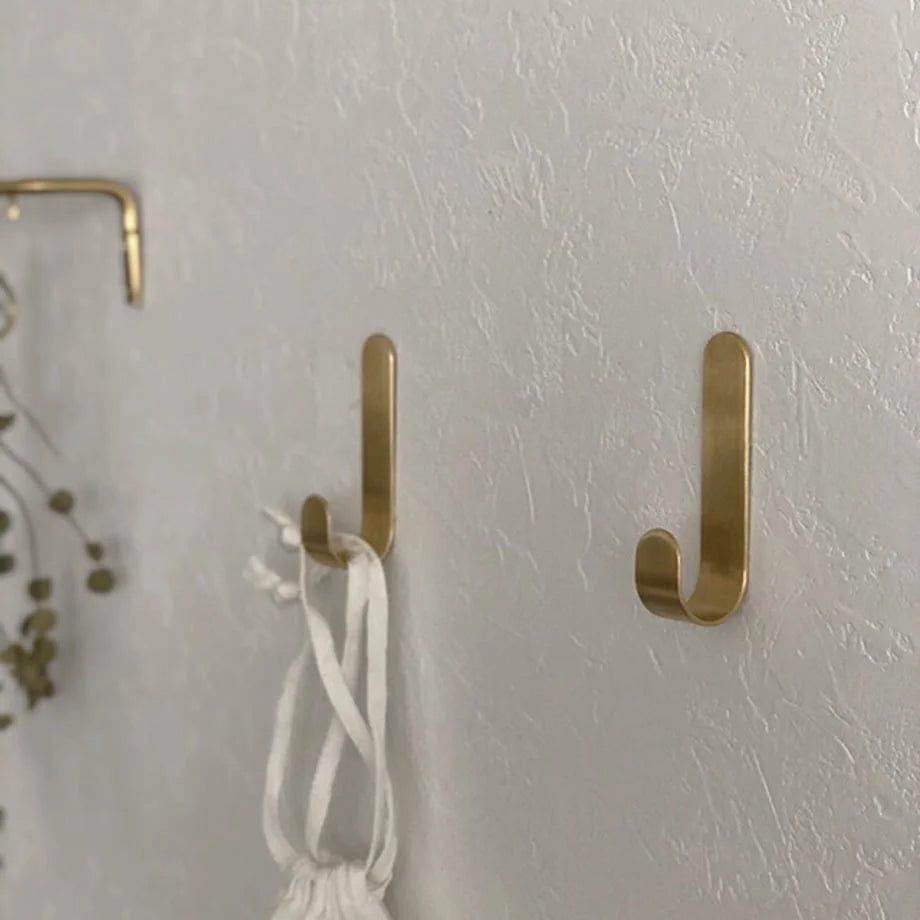 Close-up of two brushed gold J-shape metal hooks mounted on a textured white wall, one holding a white fabric pouch - RoomDen