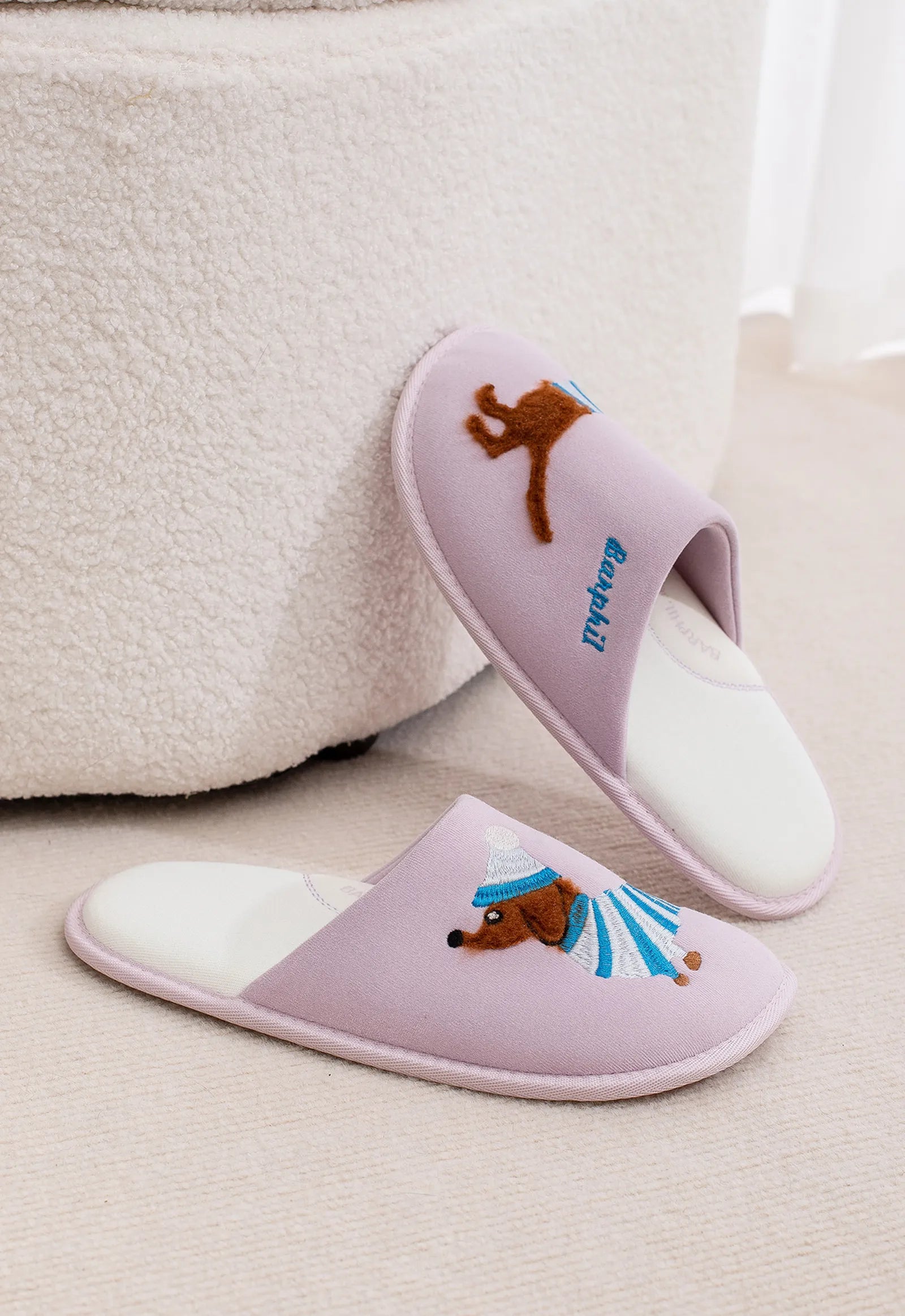 Lilac purple soft house slippers with split dog design on floor next to ottoman