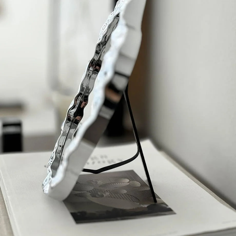 Side profile of the irregular chrome mirror, displaying its slim black stand for tabletop support - RoomDen