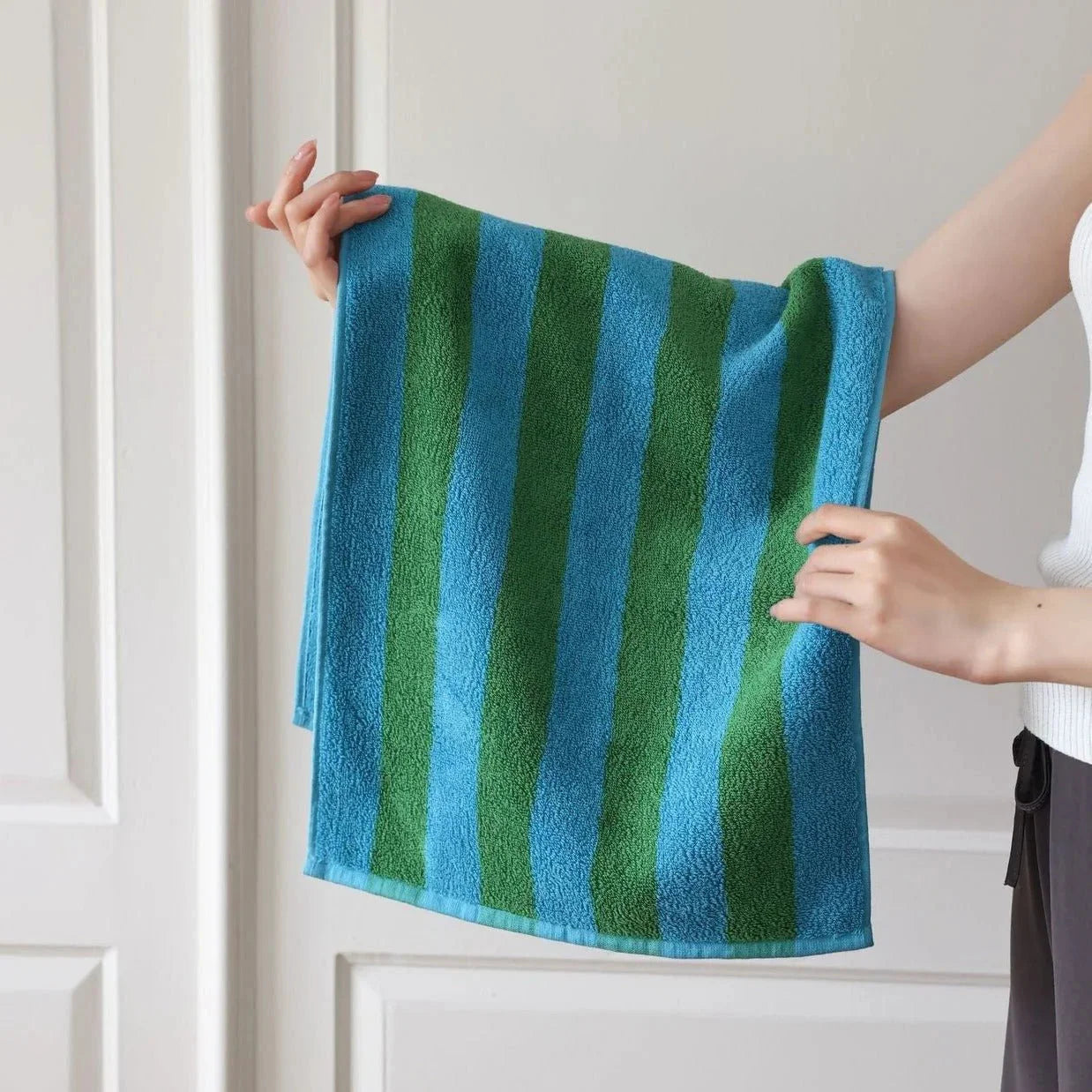A close-up of a person holding a soft, textured blue and green striped cotton face towel. - RoomDen