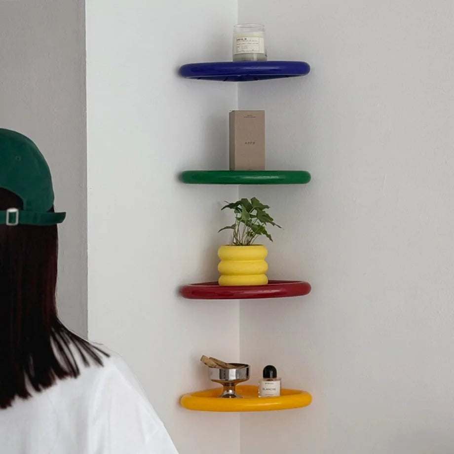 A woman looking at a set of four colorful corner shelves (blue, green, red, yellow) displaying a candle, plant, and decor. - RoomDen
