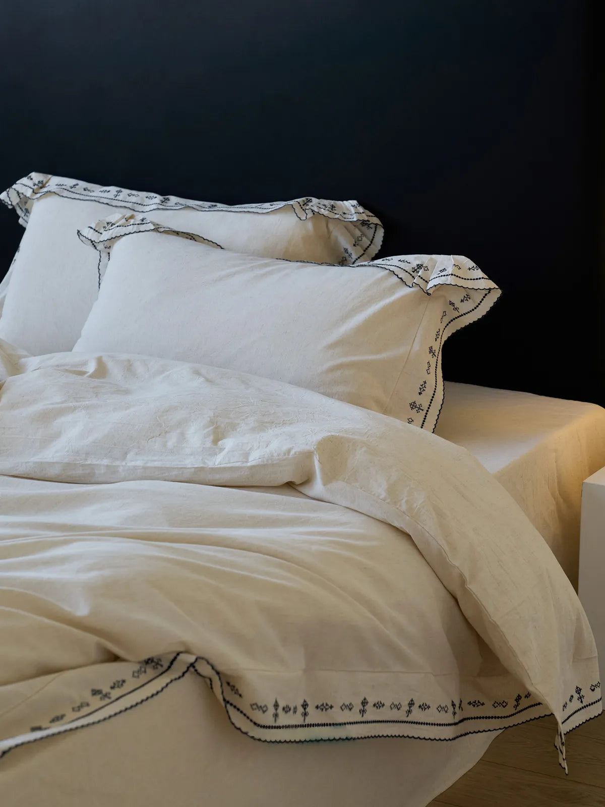 Angled shot of the Embroidered Ruffle Linen Duvet Cover and pillowcases, showing the fabric's texture against a dark background - RoomDen.