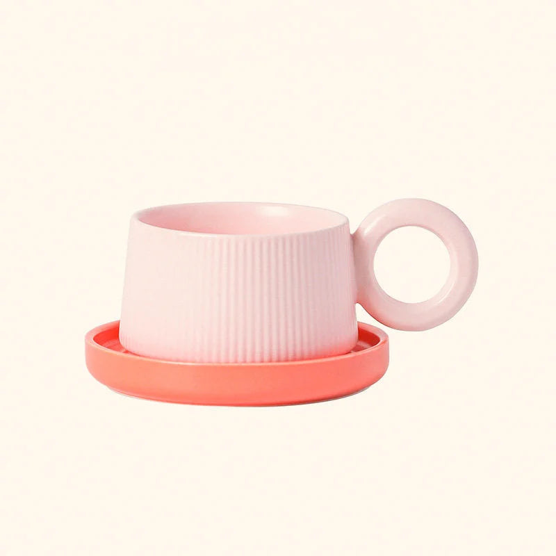 Front view of the Blush Pink ribbed ceramic mug with its round donut handle, sitting on a coral saucer. - RoomDen