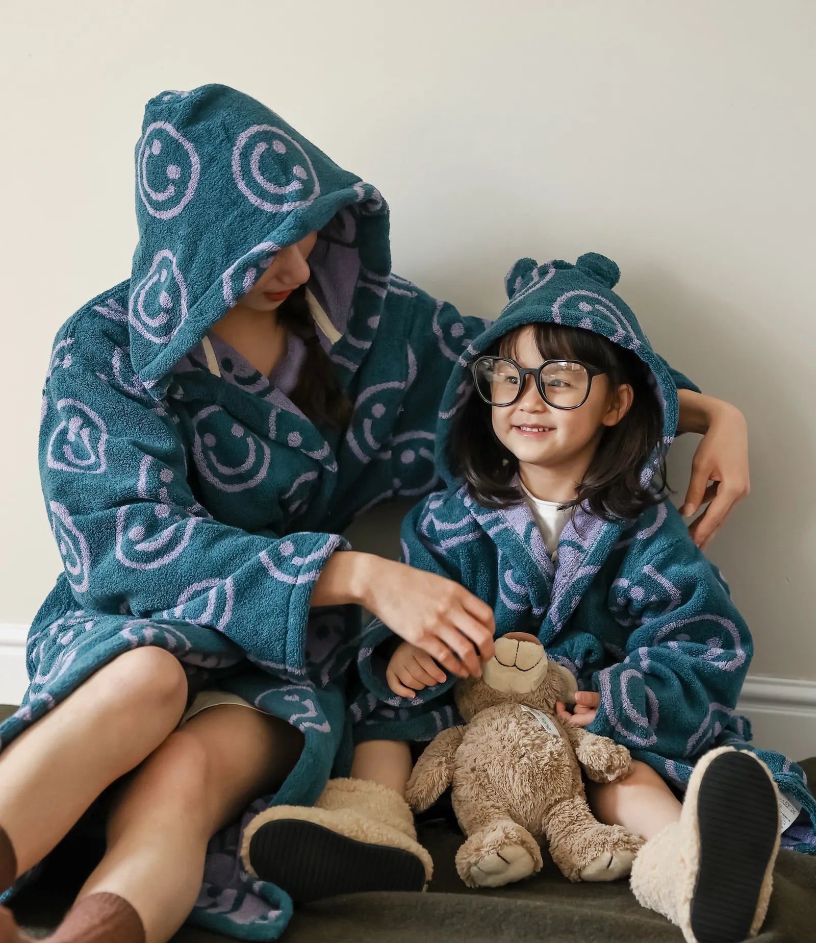 Mom and daughter sitting on the floor wearing matching teal smiley face cotton robes with hoods up