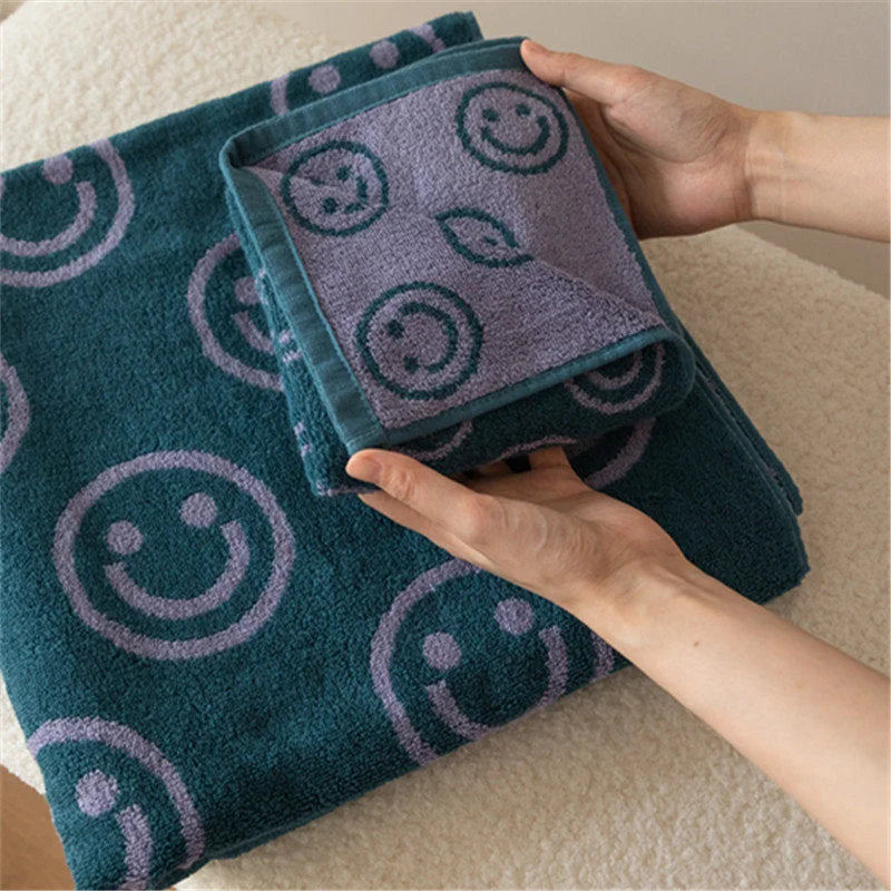 A pair of hands holding and displaying the soft texture of the lavender and teal smiley face cotton face towel over the larger bath towel - RoomDen.