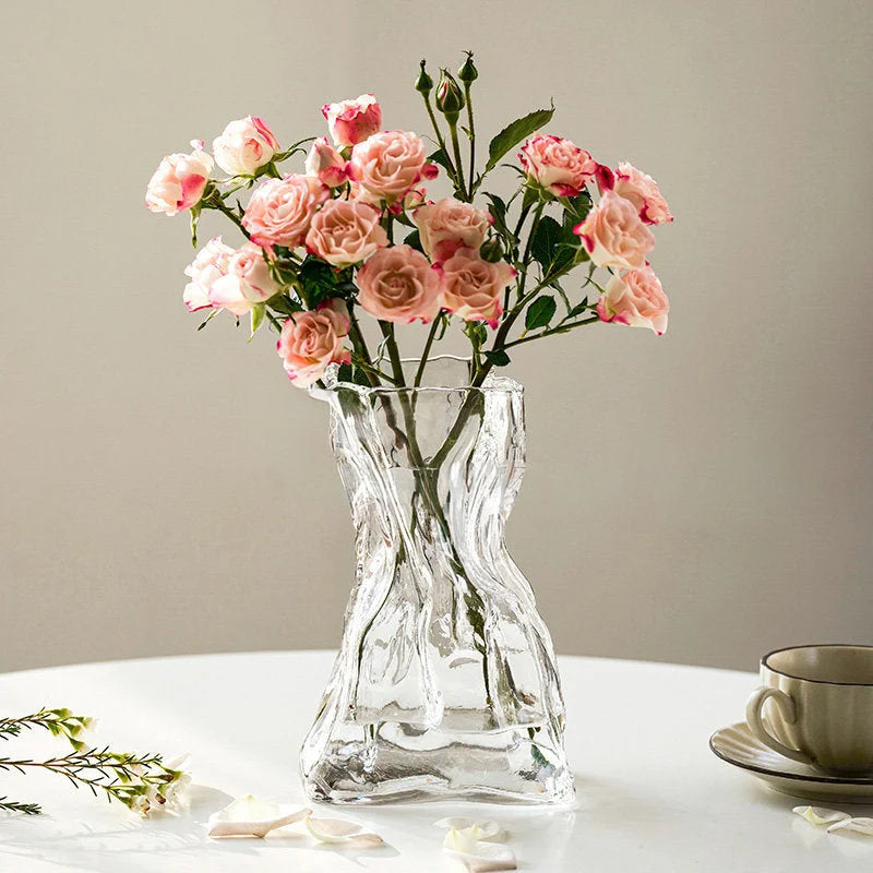 The large clear Sculptural Crumpled Glass Vase holding a bouquet of pink roses on a white table. - RoomDen