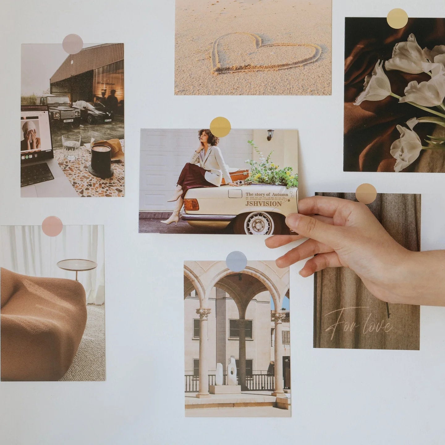 Hand arranging a minimalist wall collage kit featuring photos of architecture, flowers, and cozy interiors on a white wall. - RoomDen