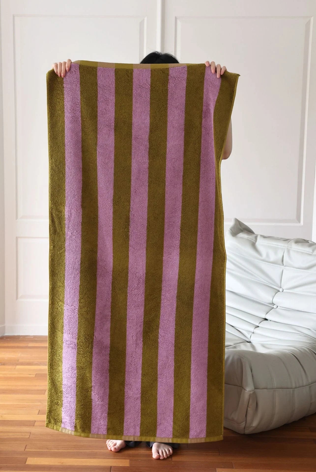 A person holding up the full-sized green and purple striped cotton bath towel, showcasing its large dimensions. - RoomDen