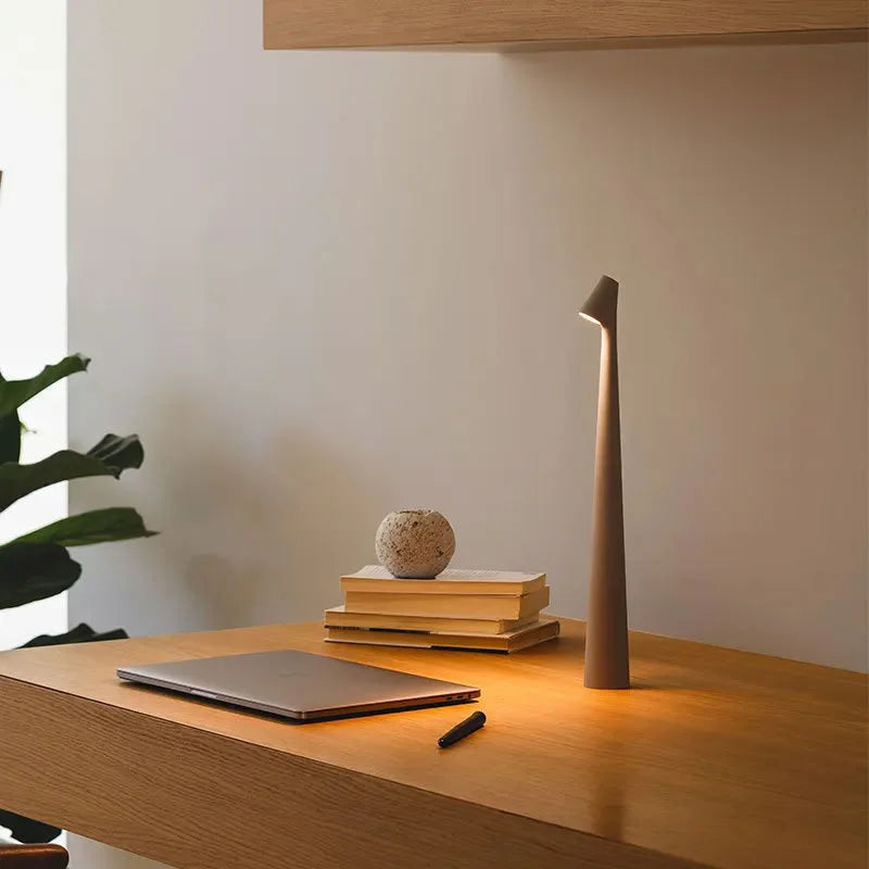 A taupe grey minimalist cordless lamp on a wooden desk next to a laptop and books, providing focused task lighting - RoomDen.