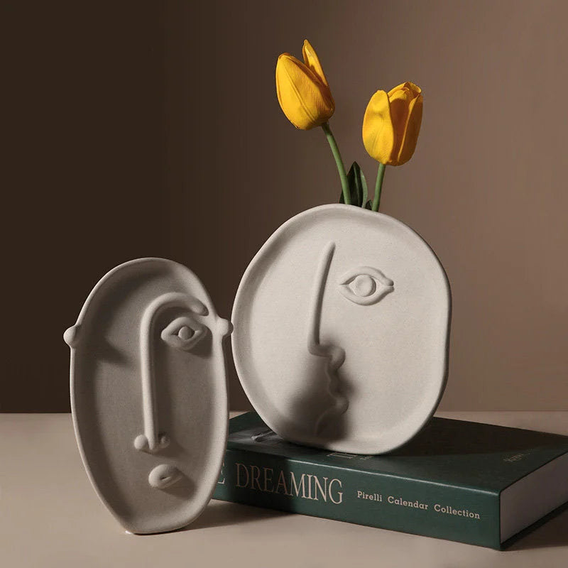 Two minimalist face vases in frosted ceramic, one holding yellow tulips, placed on books - RoomDen.