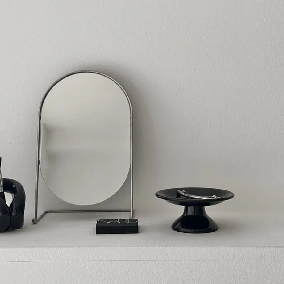 A black pedestal jewelry dish on a white vanity next to a modern silver mirror and makeup. - RoomDen
