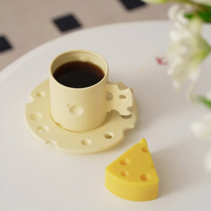 A yellow Cheese Ceramic Mug and Saucer set next to a cheese-shaped prop on a white tabletop. - RoomDen