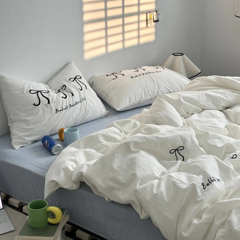 A full view of a cozy bed with a white washed cotton bedding set featuring black embroidered bows, styled in a sunlit room. - RoomDen