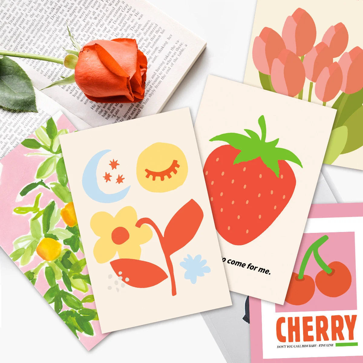Close-up detail of several cards from the wall collage kit, featuring strawberry, flower, and cherry illustrations, placed on a book. - RoomDen