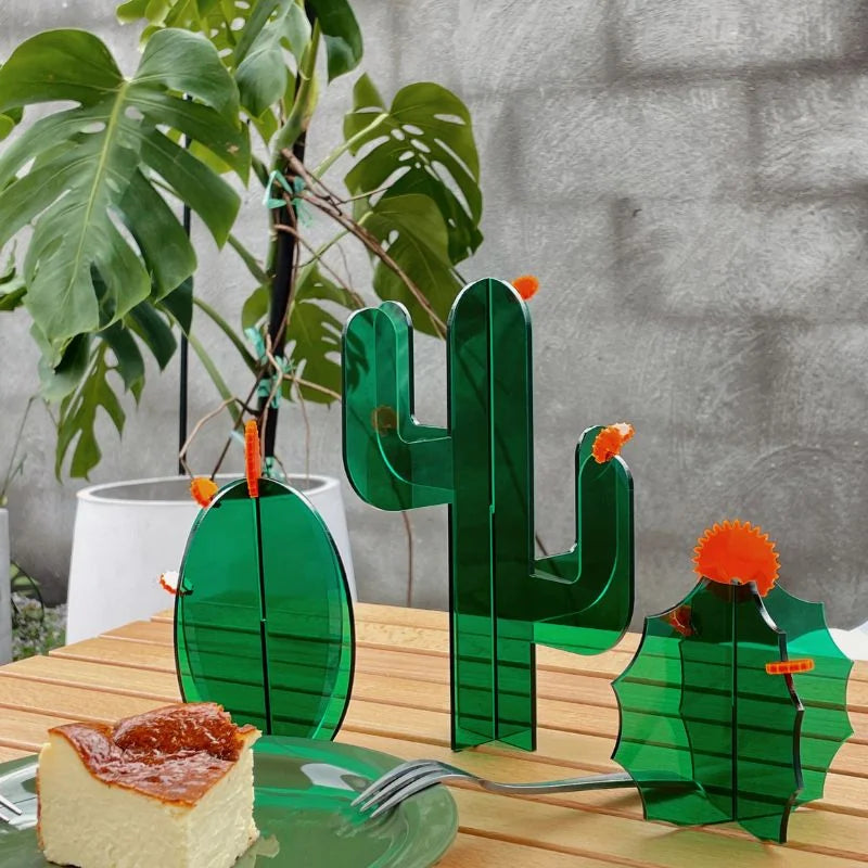 A set of three green acrylic cactus sculptures on a wooden table next to a slice of cheesecake - RoomDen