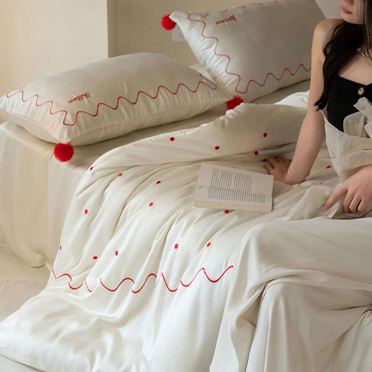 A woman sitting on a bed with a Creamy White & Red silky Tencel bedding set, showing the wavy embroidery and pom poms. - RoomDen