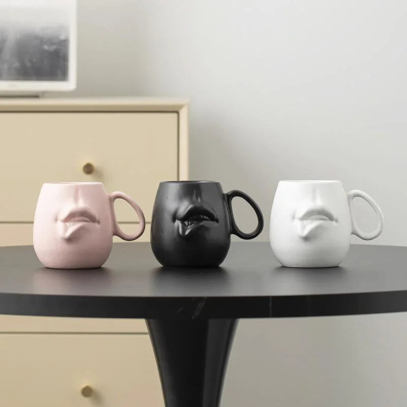The three color variants - Dusty Pink, Matte Black, and Matte White - of the abstract face mug lined up on a black table.
