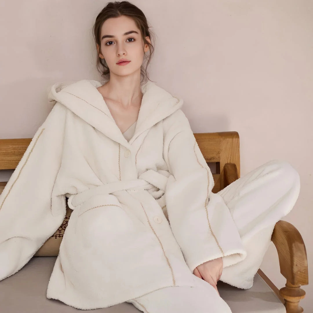 Front view of a model sitting on a bench wearing a creamy white flannel fleece pajama set with a hood and belt - RoomDen.