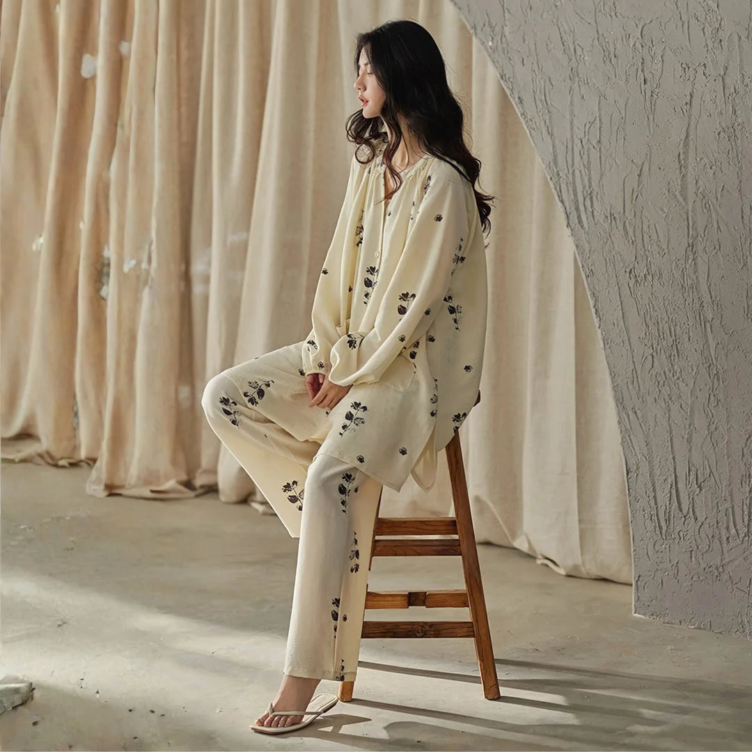 Model wearing the creamy beige botanical print pajama set, sitting sideways on a wooden stool against a textured wall - RoomDen