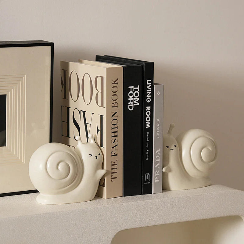 A pair of creamy white smiley snail ceramic bookends organizing books on a white shelf - RoomDen.