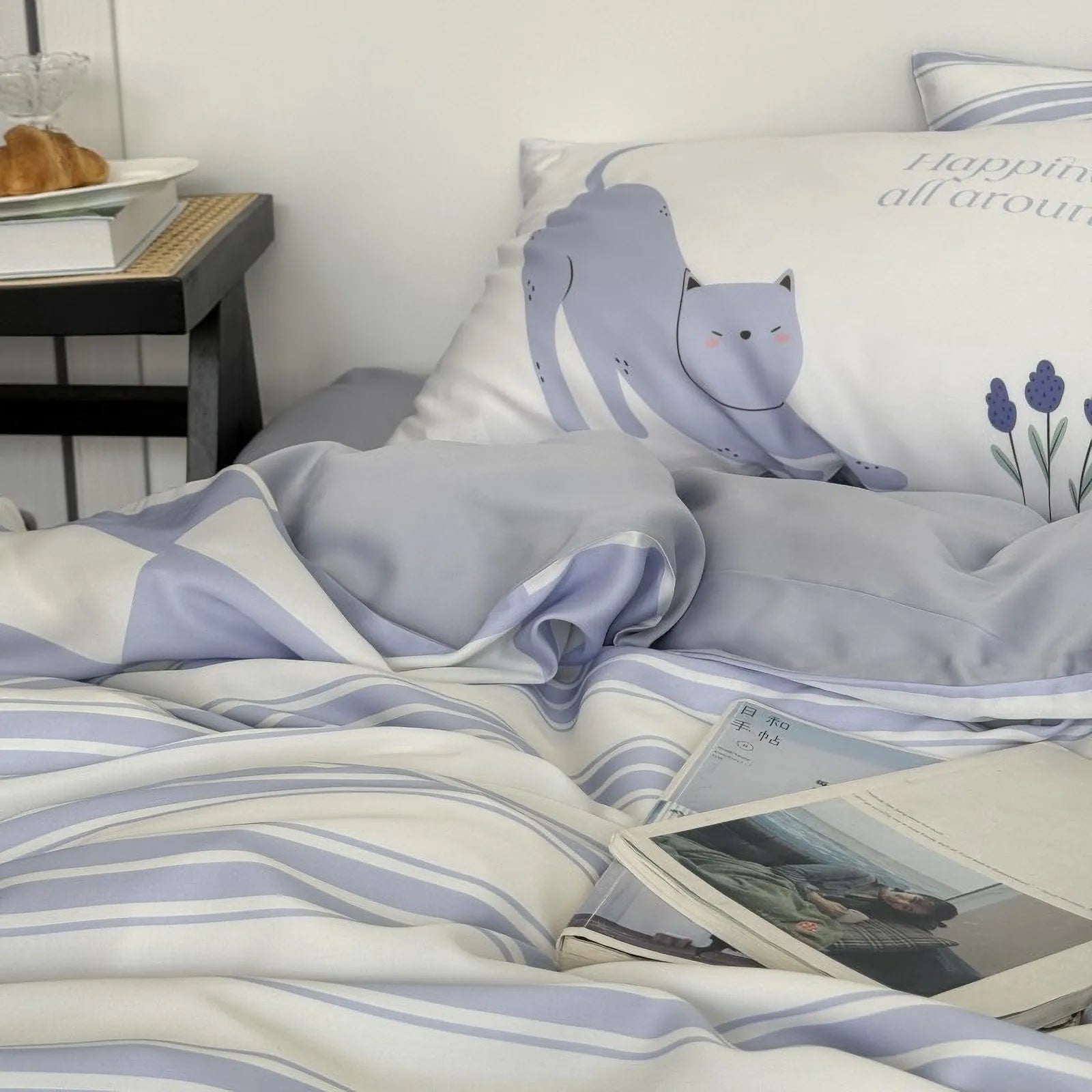 Detail shot of the cute cat illustration pillow on the Washed Cotton Bedding Set, with a book and croissant on the nightstand. - RoomDen