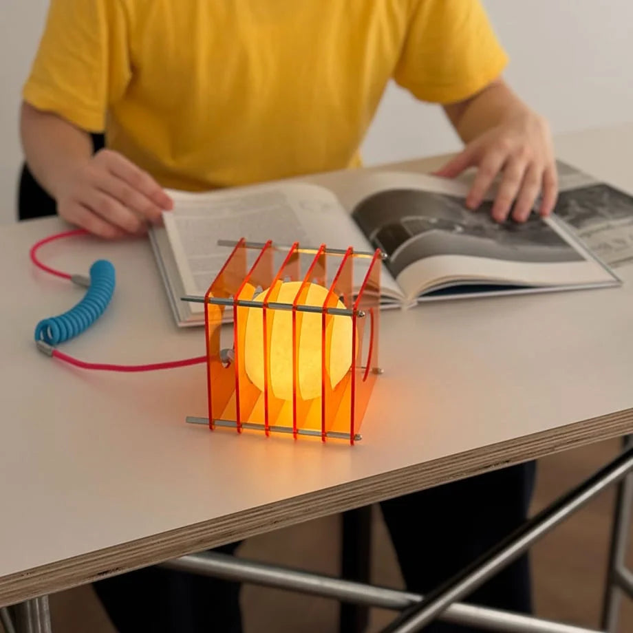 Close-up of the glowing Sunset Orange Caged Sphere Lamp while a person reads a book - RoomDen