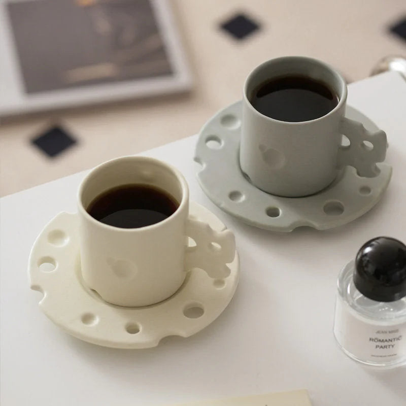 Two Cheese Ceramic Mug & Saucer sets, one in cream white and one in light grey, on a desk. - RoomDen